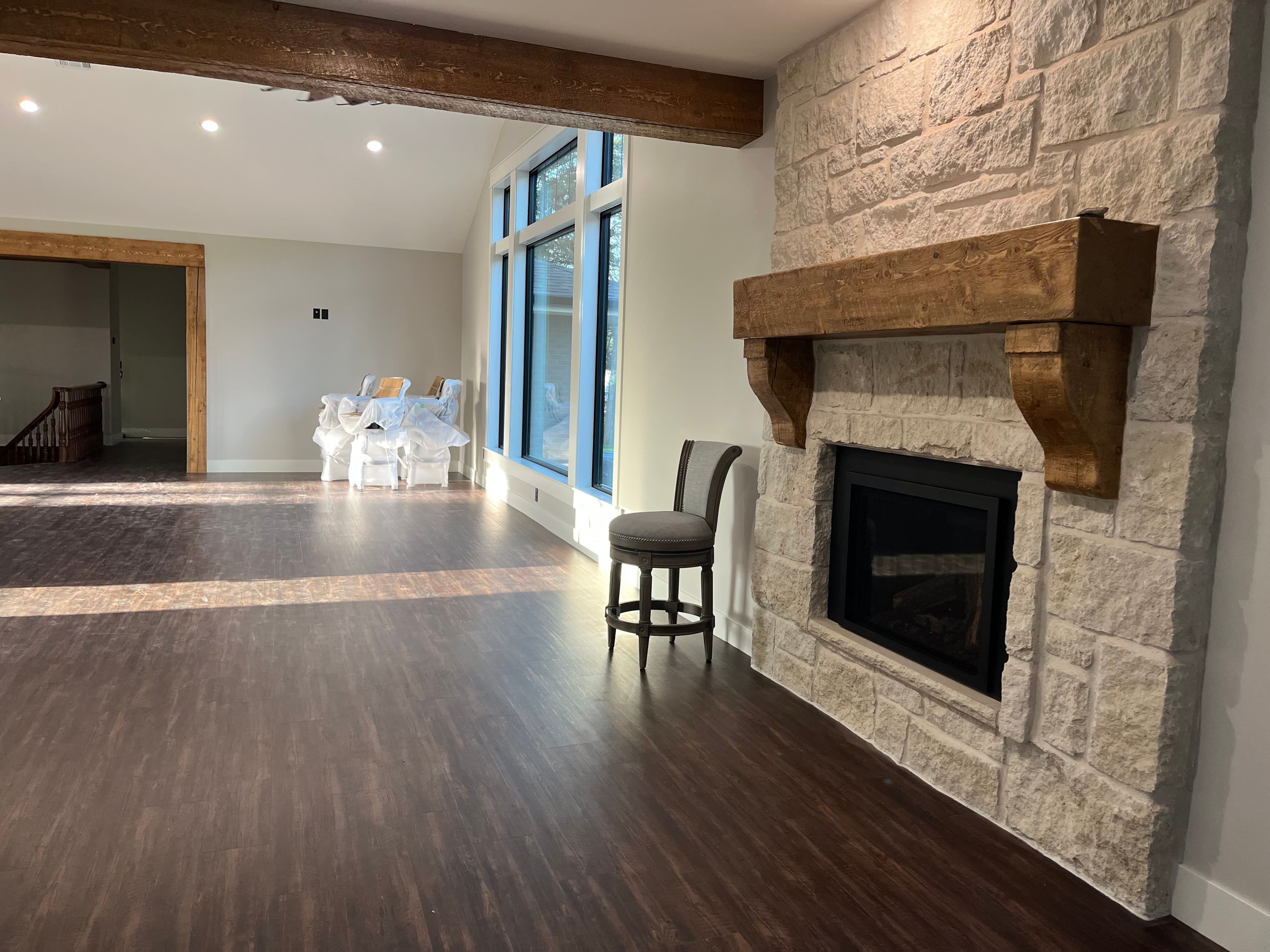 Stone fireplace with a rustic wooden mantel in a sunlit room with dark wood flooring.